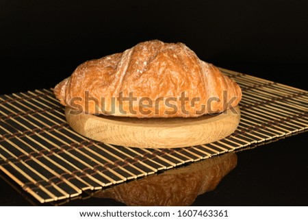 One whole sweet fresh crispy croissant on a black background lies on a brown round wooden stand, which stands on a bamboo mat.