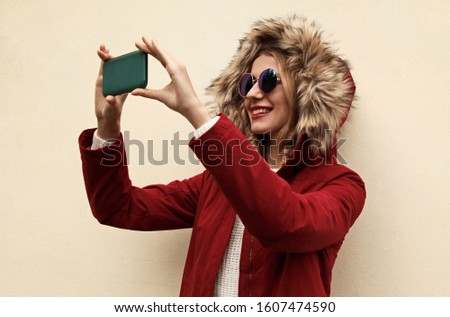Portrait of happy smiling young woman taking selfie with smartphone wearing red jacket with fur hood 