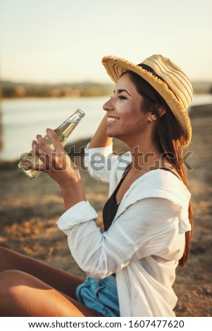 Young woman in straw hat relaxing at sunset time on the river bank and drinking beer.