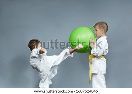 Little athlete in karategi trains kicking a green ball on a gray background