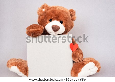 Smiling teddy bear sitting at pastel wall and holding craft card and heart Empty place for inspiration, emotional, sentimental text, lovely quote or sayings for good mood. Front view.
