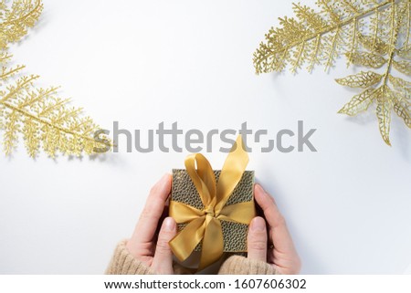 Woman in warm woolen sweater holding golden gift box on white background with decorative golden leaves. Copy space for your text. Valentine’s day background.