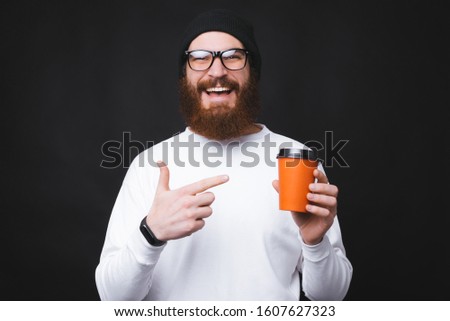 Young bearded man smiles at camera and pointing at a cup that is holding near black wall.