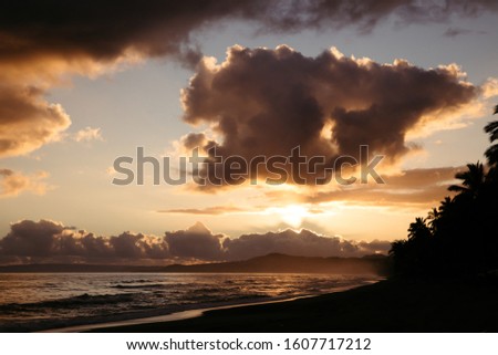 Early morning on the atlantic ocean in dominican republic. The sun rises from a mountain and  throws soft yellow light on the shore through the cloud and palm trees.