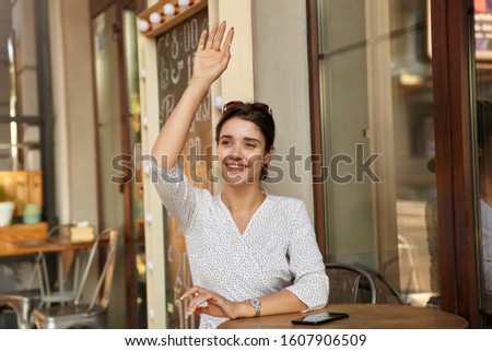 Positive young dark haired female looking aside and smiling happily while raising hand in hello gesture, meeting friends in city cafe on sunny weekend day