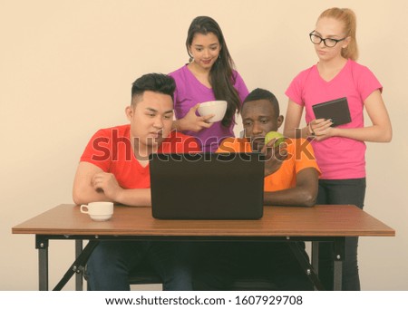 Studio shot of diverse group of multi ethnic friends using laptop while having coffee and green apple on wooden table together