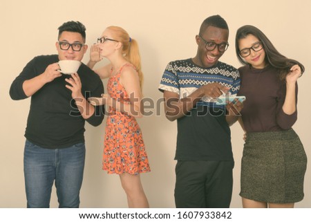 Happy diverse group of multi ethnic friends smiling while using mobile phone together with friends having coffee and whispering in the back