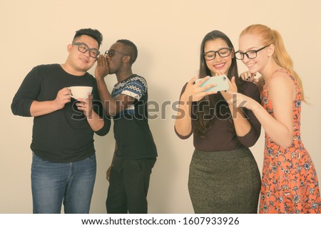 Happy diverse group of multi ethnic friends smiling while using mobile phone together with friends whispering in the back