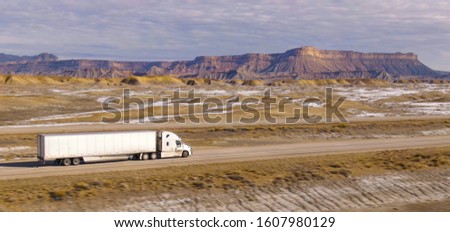 AERIAL: Semi-trailer truck speeds along the motorway running across the desert in Utah. Wintry desert landscape surrounds truck driving down the interstate highway running across rugged US wilderness. Royalty-Free Stock Photo #1607980129