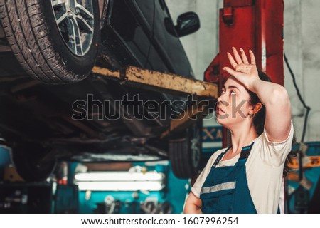 Gender equality. A young brunette woman in uniform stands near the lift with the car, and looks at the wheel, wiping the sweat from her forehead. In the background is an auto repair shop