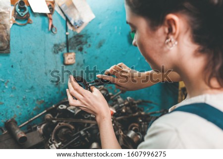 Gender equality. Portrait of a young brunette woman in uniform, who is engaged in repairing car parts. Blue wall and spare parts in the background. The view from over shoulder. Close up