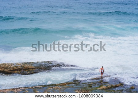Surfer man standing with surfboard on rocks looking out to the ocean, waves in the background
