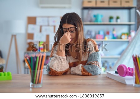 Young beautiful teacher woman wearing sweater and glasses sitting on desk at kindergarten tired rubbing nose and eyes feeling fatigue and headache. Stress and frustration concept.