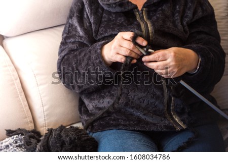 Stock photography of a woman sitting on the couch at home, knitting a gray and black scarf. Close up photography focus on the hands.