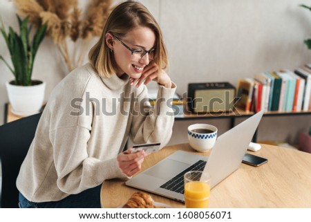 Photo of joyful blonde woman holding credit card and using laptop while sitting at table in cozy living room