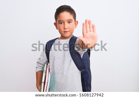 Beautiful student kid boy wearing backpack holding books over isolated white background with open hand doing stop sign with serious and confident expression, defense gesture