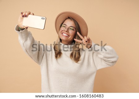 Portrait of funny young woman in hat gesturing peace sign and taking selfie photo on cellphone isolated over beige background