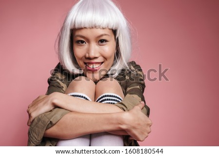 Image of positive asian girl wearing white wig smiling and hugging her knees isolated over pink background