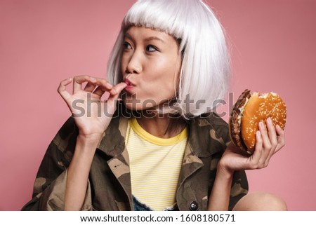 Image of young asian girl wearing white wig having fun and eating big burger isolated over pink background