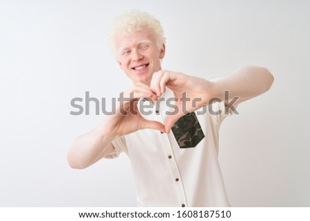 Young albino blond man wearing casual shirt standing over isolated white background smiling in love showing heart symbol and shape with hands. Romantic concept.