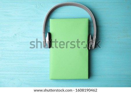 Book with blank cover and headphones on light blue wooden background, flat lay