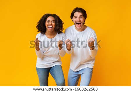 Big Win. Cheeful mixed-race couple celebrating success, raising hands with excitement over yellow background in studio, free space