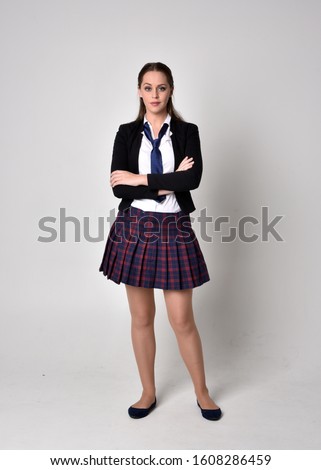 full length portrait of a pretty brunette girl wearing a school uniform of black jacket and plaid skirt. Standing pose  against a studio background.