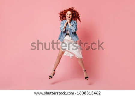 Full length view of amazed ginger lady jumping on pink background. Studio shot of active curly girl in denim jacket and high-heeled shoes.