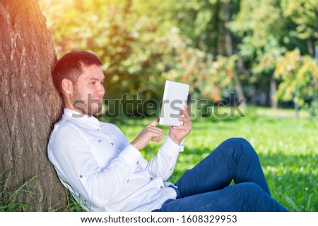 Man using tablet computer under tree in park on sunny day. Handsome guy looking at tablet screen. Happy smiling man sitting on green grass leaning against tree. Digital technology and communication