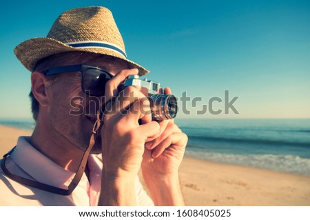 Retro tourist photographer with hat and sunglasses taking a photo with an old fashioned point and shoot camera on an empty beach