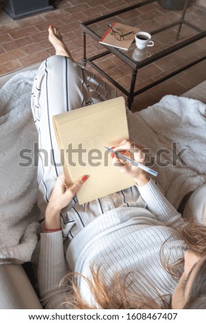 Young woman starting to write in a blank notebook sitting on the sofa at home with a pen. The notebook is blank.