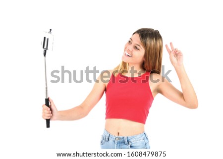 Cute teenager girl doing a two finger gesture with her hand while holding a selfie stick with her mobile phone against a white background