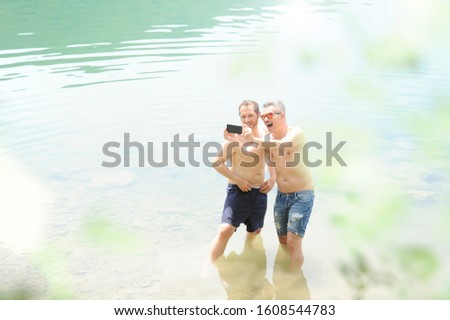 Two men standing together outdoors taking a selfie and smiling. Best friends taking a selfie while standing into the water of the lake on a summer day. Men wearing only jeans shorts. Bare-chested.