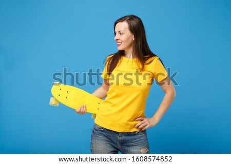 Beautiful young brunette woman girl in yellow t-shirt posing isolated on blue wall background in studio. People sincere emotions lifestyle concept. Mock up copy space. Hold skateboard, looking aside