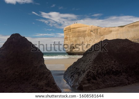 Tunnel Beach in Otago, New Zealand. The sandstone headland at the end of the beach with two tourists on top is in the background, viewed between two large, silhouetted rocks in the foreground.