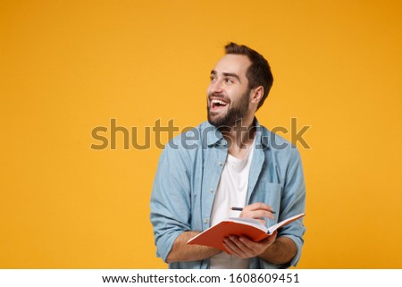 Cheerful young student man in casual blue shirt posing isolated on yellow orange wall background, studio portrait. People lifestyle concept. Mock up copy space. Writing note in notebook looking aside