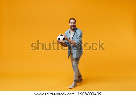 Excited cheerful young man in casual blue shirt posing isolated on yellow orange wall background, studio portrait. People sincere emotions lifestyle concept. Mock up copy space. Holding soccer ball