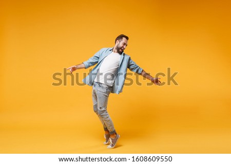 Laughing young bearded man in casual blue shirt posing isolated on yellow orange background studio portrait. People emotions lifestyle concept. Mock up copy space. Standing on toes, spreading hands