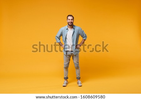 Cheerful young bearded man in casual blue shirt posing isolated on yellow orange background, studio portrait. People emotions lifestyle concept. Mock up copy space. Standing with arms akimbo on waist Royalty-Free Stock Photo #1608609580