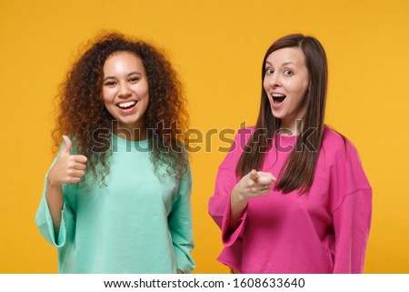 Two women friends european african american in pink green clothes posing isolated on yellow background. People lifestyle concept. Mock up copy space. Showing thumb up, pointing index finger on camera