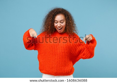 Amazed young african american girl in casual orange knitted clothes isolated on pastel blue wall background studio portrait. People lifestyle concept. Mock up copy space. Pointing index fingers down