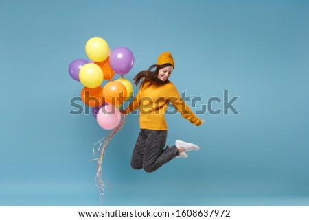 Smiling young woman girl in sweater and hat posing isolated on blue background. Birthday holiday party, people emotions concept. Mock up copy space. Celebrating holding colorful air balloons jumping