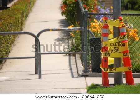A caution tape on bars in front of the fences of a park covered in greenery under sunlight