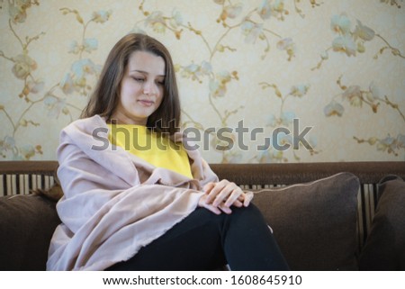 A picture of nice and thoughtful girl sits at sofa and reads a book. She has brown blanket on her shoulders. Woman got cold. She suts at window