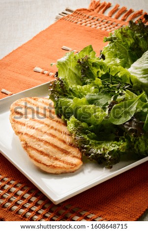 Appealing grilled chicken fillet served on a white square plate with leaf salad aside, on top of a orange table mat.