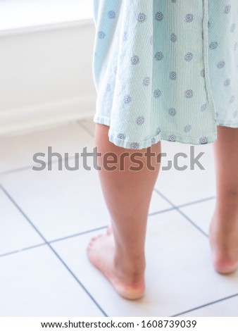 A closeup photograph of a middle aged mixed race African American woman's legs as she is standing on a cold tile floor barefoot wearing a blue patterned hospital gown or robe staring out the window.