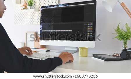 Side shot of young man while using 
 Video editor on the work desk. Potted plant, coffee cup, lamp, pencil holder, computer, on working desk.