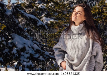 Portrait of a young beautiful brunette woman with blue eyes and freckles on face in winter snowy mountain landscape. Woman dreesed in sweater standing next to fur tree. Copy space