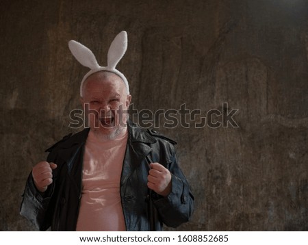 elderly man in pink pajamas, rabbit ears and leather coat of exhibition maniac is dancing against background of imitating mold. concept of perversions of bizarre people. Gray beard and wrinkled face