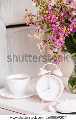 Alarm clock closeup, a cup of coffee and bouquet of pink flowers background in the morning sunlight